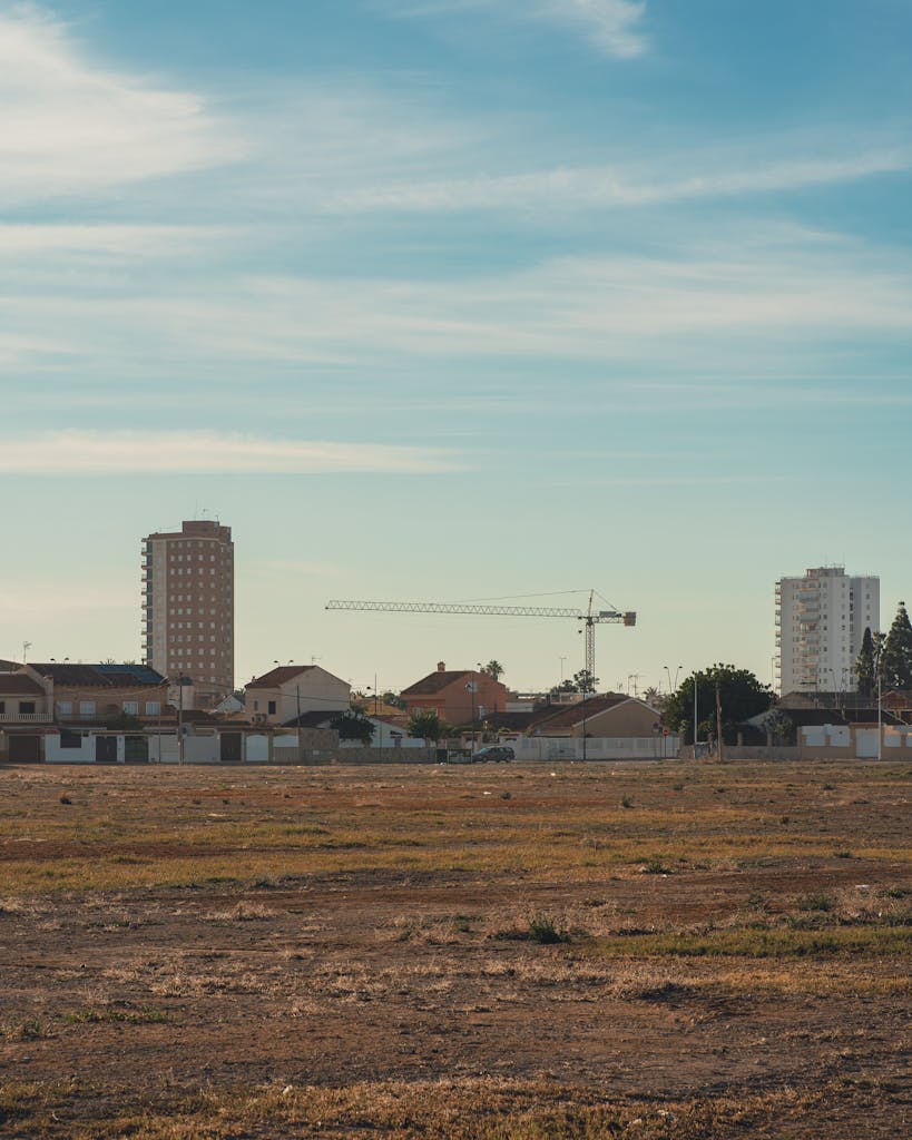 A cityscape featuring buildings, open land, and a visible construction crane under a blue sky.
